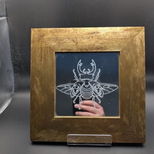 Puede incluir: Espejo cuadrado enmarcado en dorado, con un diseño detallado de insecto blanco grabado en la superficie reflectante. El marco tiene un aspecto texturizado. El espejo se exhibe sobre un soporte de plástico transparente.