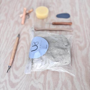 Peut inclure: Un ensemble d'outils de poterie et d'argile. L'ensemble comprend un sac d'argile grise, une éponge, un outil à manche en bois et d'autres outils de sculpture. Une étiquette bleue avec le texte "na ART & DESIGN" est attachée au sac d'argile.