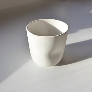 Könnte beinhalten: Eine weiße Keramiktasse mit strukturierter Oberfläche. Die Tasse hat einen abgerundeten Boden und einen leicht ausgestellten Rand. Die Tasse ist etwa 10 cm hoch und 8 cm breit.