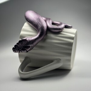 Peut inclure: Une tasse en céramique blanche avec une surface texturée, ornée d'un serpent violet décoratif enroulé sur le bord. Le serpent, avec une texture d'écailles détaillée, semble se reposer sur la tasse.