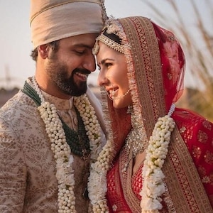 May include: A couple in traditional Indian wedding attire, the groom wearing a beige sherwani with a white floral garland and the bride wearing a red lehenga with a white floral garland. They are holding hands and looking at each other.