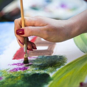 May include: A wooden-handled paintbrush with metal ferrule applying paint to a canvas. The canvas features textured strokes of green, purple, and red pigment, with a brown rectangular shape visible in the background of the painting.
