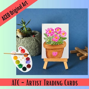 May include: A small watercolor painting on a miniature wooden easel depicting a brown terracotta pot with pink flowers and a heart detail. A white plastic paint palette with various colors and a thin paintbrush are positioned nearby.
