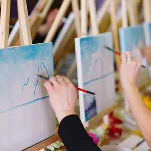 May include: Multiple wooden easels holding white canvases with light blue sky and mountain landscape paintings in progress. Each canvas features a horizontal blue line and mountain outlines, with paintbrushes applying blue paint to the surfaces.