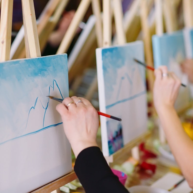 May include: Multiple wooden easels holding white canvases with light blue sky and mountain landscape paintings in progress. Each canvas features a horizontal blue line and mountain outlines, with paintbrushes applying blue paint to the surfaces.