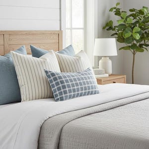May include: A bed with a light wood headboard and a white duvet. The bed is styled with several pillows in shades of blue and cream, including a checkered pillow. A grey textured blanket is at the foot of the bed.