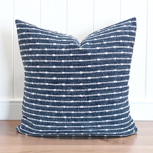 Peut inclure: Un coussin carré décoratif avec un fond bleu marine et des lignes blanches pointillées horizontales. Le coussin est fait d'un tissu texturé et est posé sur une surface en bois. C'est un article de décoration intérieure.