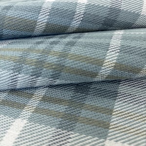 May include: A woven fabric featuring a plaid pattern in shades of light blue, slate grey, and white. The textile displays a textured surface with intersecting horizontal and vertical lines that create a classic checkered design across the material.