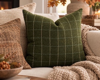May include: A square throw pillow in a deep olive green fabric featuring a subtle white grid pattern. The pillow has a soft, textured appearance and is positioned among other neutral-toned cushions and a chunky knit throw blanket.