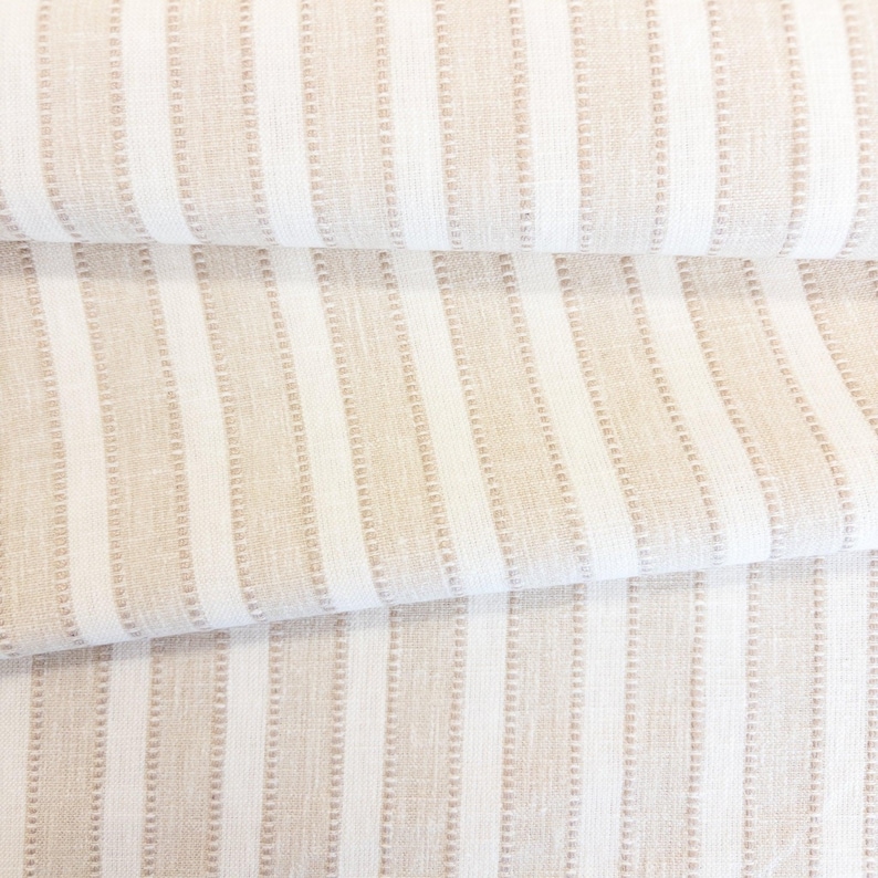 May include: A folded piece of fabric featuring a vertical striped pattern in alternating shades of beige and white. The design consists of solid white stripes paired with textured beige stripes that have a subtle, repeating dotted line detail.