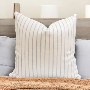 May include: A square decorative pillow with a white background and vertical blue stripes. The pillow is placed on a bed with a light blue pillow and a brown blanket. The pillow has a soft, textured appearance.