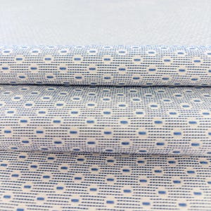 Puede incluir: Tela enrollada con un patrón repetitivo de pequeños rectángulos y líneas azules sobre un fondo blanco. La tela está tejida de forma apretada, con una apariencia texturizada. El patrón crea un diseño geométrico, adecuado para varios proyectos textiles.