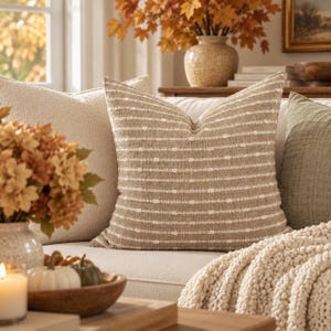May include: A square throw cushion featuring a tan and white horizontal striped pattern with a textured woven fabric. The cushion is positioned on a sofa alongside a solid cream cushion and a solid olive green cushion, creating a layered look.