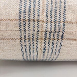 Op de afbeelding: Een close-up van een geweven stof met een ruitpatroon. Het textiel heeft een cr&egrave;mekleurige basis met verticale en horizontale strepen in donkerblauw en lichtbruin, wat een rasterontwerp cre&euml;ert met een zichtbare, getextureerde weving.