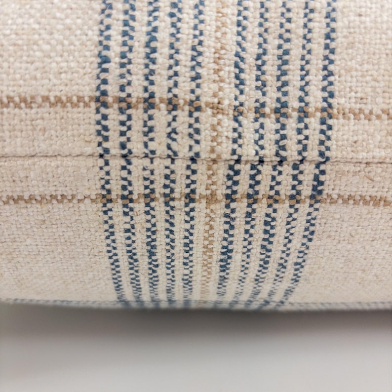 Op de afbeelding: Een close-up van een geweven stof met een ruitpatroon. Het textiel heeft een cr&egrave;mekleurige basis met verticale en horizontale strepen in donkerblauw en lichtbruin, wat een rasterontwerp cre&euml;ert met een zichtbare, getextureerde weving.