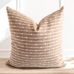 May include: A square cushion featuring a tan and white striped design. The fabric has a textured appearance with horizontal stripes and small white dots. The cushion is placed on a wooden surface, against a white wall.