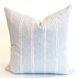 May include: A square throw cushion featuring a light blue and white striped pattern. The design consists of thin vertical blue lines alternating with wider white bands, accented by a central dotted line within each blue stripe across the fabric.