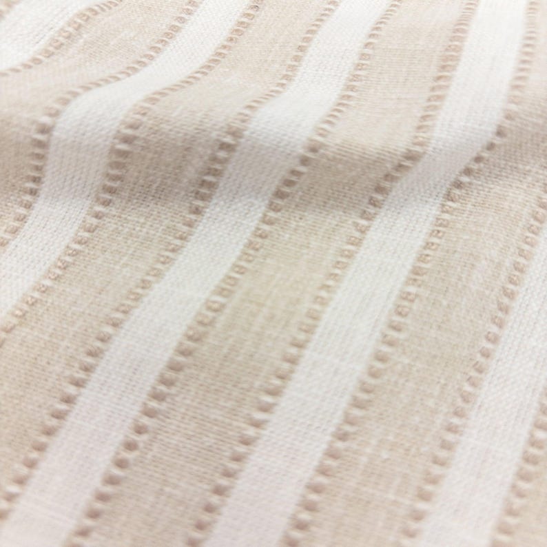 May include: A woven fabric featuring a repeating pattern of vertical stripes in alternating shades of beige and white. The texture includes subtle raised dotted lines running parallel to the stripes, creating a tactile surface with a soft, natural appearance.