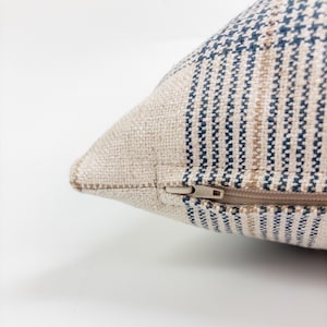 Op de afbeelding: Een kussenhoes met textuur gemaakt van geweven beige stof met verticale strepen in marineblauw en lichtbruin. De hoek van het kussen toont een verborgen ritssluiting met een kleine, lichtgekleurde plastic ritstrekker ge&iuml;ntegreerd in de naad.