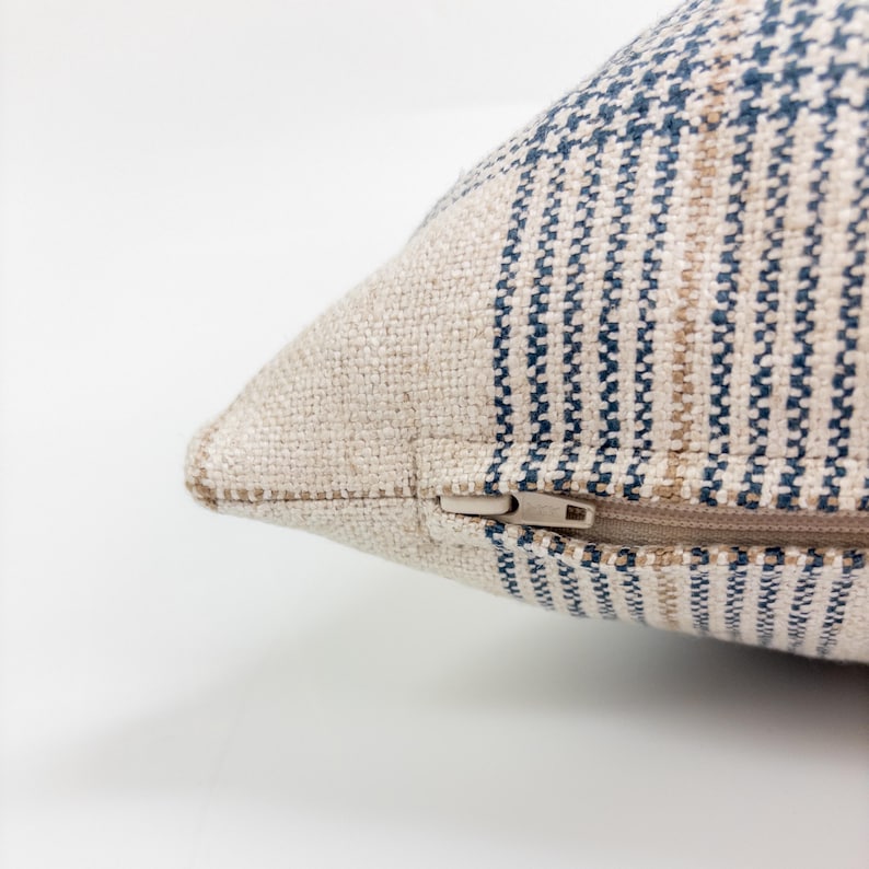 Op de afbeelding: Een kussenhoes met textuur gemaakt van geweven beige stof met verticale strepen in marineblauw en lichtbruin. De hoek van het kussen toont een verborgen ritssluiting met een kleine, lichtgekleurde plastic ritstrekker ge&iuml;ntegreerd in de naad.