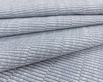 Op de afbeelding: Een gevouwen stuk stof met een geweven geometrisch patroon in tinten lichtblauw en wit. Het textiel vertoont een herhalend gestructureerd rasterontwerp met afwisselende verticale en horizontale lijnen, wat een subtiel dambordeffect creëert.