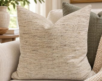 May include: A square throw pillow featuring a textured, woven fabric in a neutral beige and cream color palette with subtle dark brown flecks throughout the material. The pillow has a soft, slightly rumpled appearance and a knife-edge finish.