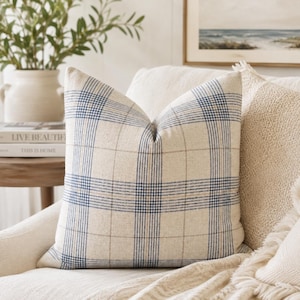 Op de afbeelding: Een vierkant sierkussen van beige linnen-achtig materiaal met een blauw en beige ruitpatroon. Het ontwerp bestaat uit elkaar kruisende verticale en horizontale lijnen die een raster vormen op het oppervlak van de kussenhoes.