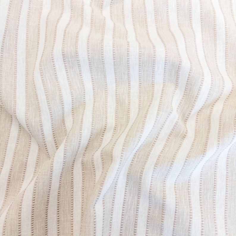 May include: A fabric featuring a vertical striped pattern in alternating shades of white and light beige. The material has a textured weave with subtle, raised linear details running parallel throughout the length of the cloth, creating a soft, draped appearance.