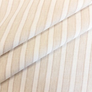 May include: A folded piece of fabric featuring a repeating pattern of vertical stripes in alternating shades of beige and white. The texture of the material appears woven with a subtle grid-like detail running along the length of the stripes.