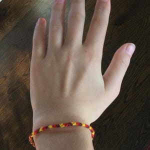 May include: A close-up of a person's wrist wearing a friendship bracelet with red, yellow, and brown threads.