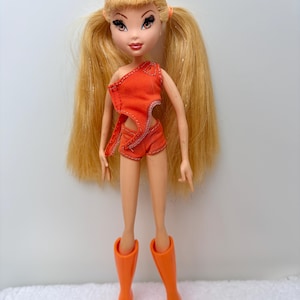 Op de afbeelding: Pop met lang blond haar in knotjes, gekleed in een oranje outfit en bijpassende kniehoge laarzen. De pop heeft een lichte huidskleur, met gedetailleerde gelaatstrekken, waaronder grote ogen en rode lippen. De pop staat op een witte ondergrond.