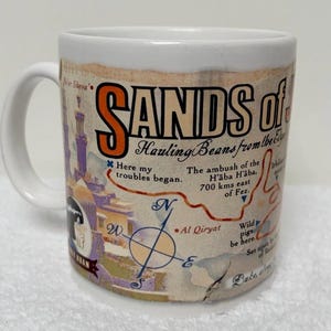 Op de afbeelding: Een witte keramische mok met een zwart handvat en de tekst "But First, Coffee" in een handschriftstijl. De mok staat op een wit oppervlak. De mok is ongeveer 10 cm hoog.