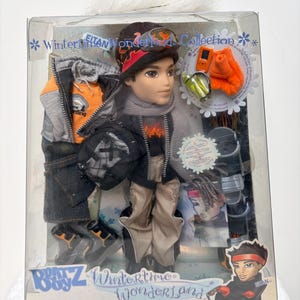 Könnte beinhalten: Eine Bratz Wintertime Wonderland Puppe in einer transparenten Plastikbox. Die Puppe trägt Winterkleidung, darunter eine schwarze Jacke, eine beige Hose und ein rot-schwarzes Stirnband. Zubehör umfasst orangefarbene Handschuhe und eine kleine grüne Flasche. Auf der Box steht "Wintertime Wonderland Collection".