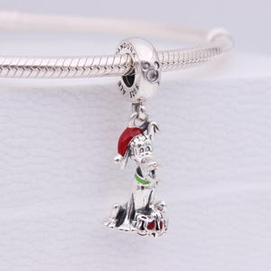 May include: A silver charm featuring a cartoon dog wearing a red Santa hat and a green collar. The charm is attached to a silver bracelet. The dog's paws are red. The charm is a festive accessory.