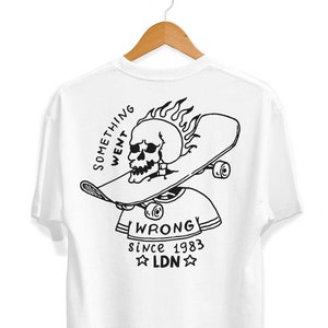 Op de afbeelding: Wit T-shirt met een zwart-witte grafische print van een schedel op een skateboard met vlammen. De tekst "SOMETHING WENT WRONG! since 1983 LDN" is onder de print gedrukt.