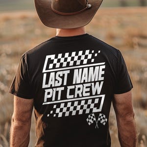 Op de afbeelding: Zwart t-shirt met de witte tekst "LAST NAME PIT CREW" en geblokte vlagafbeeldingen. De persoon draagt een bruine cowboyhoed.