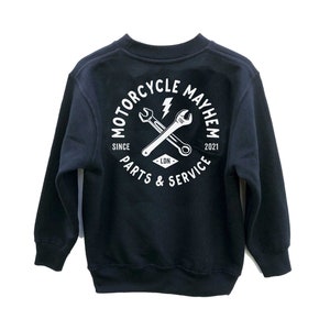 Könnte beinhalten: Ein schwarzes Sweatshirt mit einem weißen Kreis-Motiv auf der Rückseite. Das Motiv zeigt den Text "Motorcycle Mayhem" mit einem Schraubenschlüssel und einem Schraubendreher, die sich im Kreis kreuzen. Der Text "Since 2021" und "Parts & Service" ist ebenfalls im Motiv enthalten.