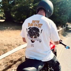 May include: White t-shirt with a black panther graphic and the text "Bite the Dust" in orange. The panther has a large mouth with sharp teeth and is surrounded by a banner that reads "Est. Premium Vintage 1942".