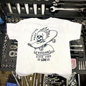 K&ouml;nnte beinhalten: Wei&szlig;es T-Shirt mit einer schwarz-wei&szlig;en Grafik eines Sch&auml;dels auf einem Skateboard mit Flammen. Der Text "Something Went Wrong" befindet sich &uuml;ber dem Sch&auml;del und "Since 1983 LDN" unter dem Sch&auml;del.