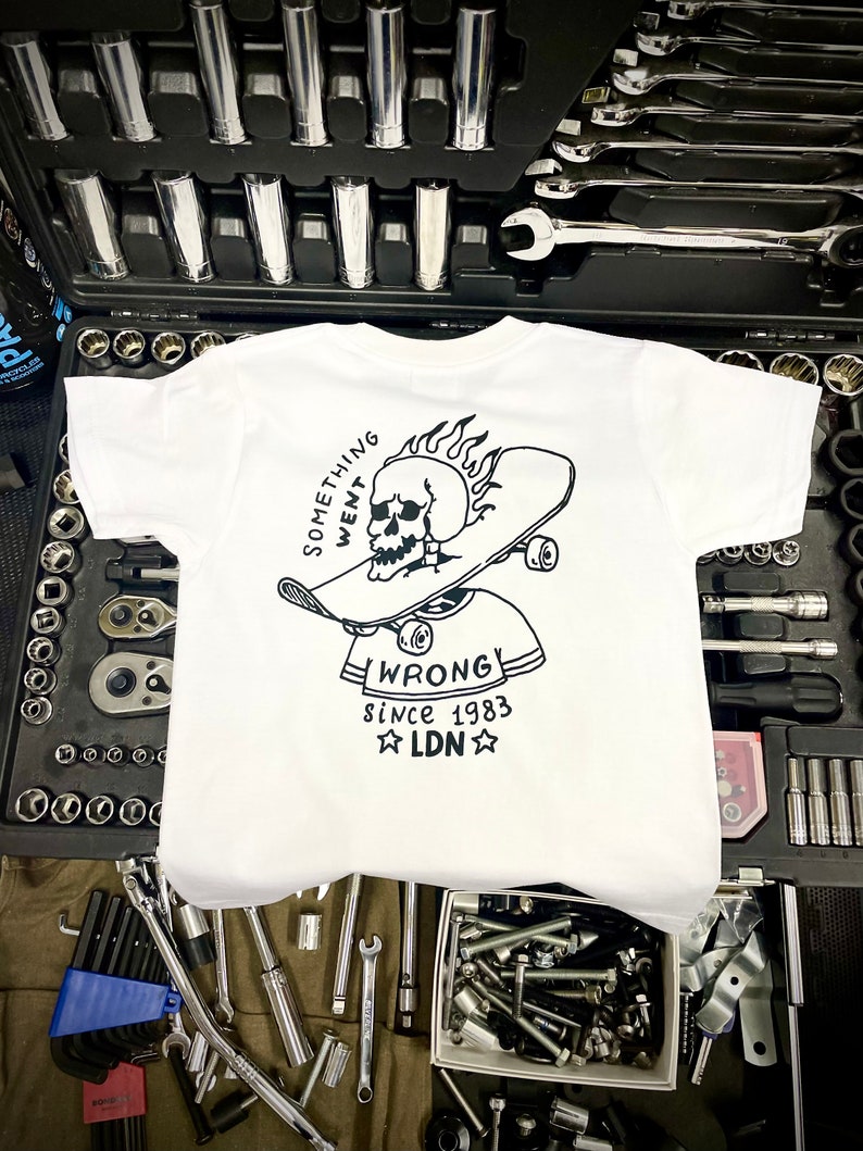K&ouml;nnte beinhalten: Wei&szlig;es T-Shirt mit einer schwarz-wei&szlig;en Grafik eines Sch&auml;dels auf einem Skateboard mit Flammen. Der Text "Something Went Wrong" befindet sich &uuml;ber dem Sch&auml;del und "Since 1983 LDN" unter dem Sch&auml;del.