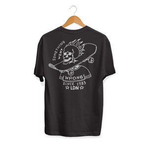 K&ouml;nnte beinhalten: Schwarzes T-Shirt mit einem wei&szlig;en Grafikdruck eines Sch&auml;dels, der ein Skateboard mit Flammen tr&auml;gt. Der Text "Something Went Wrong" und "Since 1983 LDN" ist unter dem Grafikdruck aufgedruckt.