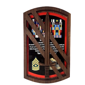 May include: A wooden plaque with a dark brown frame and a red inner border. The plaque features a central sword design and displays military insignia, ribbons, and text. The text includes phrases like "On behalf of" and "thank you".