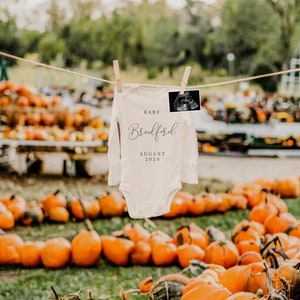 May include: A white onesie with the text "Baby Bradford August 2024" hanging on a clothesline above a pile of orange pumpkins.