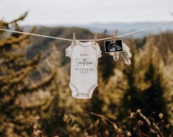 以下が含まれることがあります： 「Baby Smithson December 2023」と書かれた白いベビーロンパースが洗濯ロープにかけられています。隣には白黒の超音波画像がかけられています。