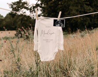 May include: A white baby onesie with the text "BABY Madison FEBRUARY 2024" hanging on a clothesline in a field of long grass. A black and white ultrasound image is attached to the onesie.
