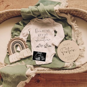 May include: A white baby onesie with the text "Our next great adventure" and a mountain range graphic. A black and white ultrasound image is placed on top of the onesie. A wooden sign with the text "Baby Fontain August 2024" is in the image. A green blanket and a rainbow-colored woven wall hanging are also in the image.