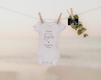 May include: A white baby onesie with the text "Baby Taylor" and "March 2024" printed on it. The onesie is hanging on a clothesline with a small ultrasound image hanging next to it.