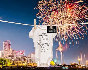 May include: A white onesie with the text "countdown to baby Reynolds" and "August 2024" hanging on a clothesline against a backdrop of fireworks over a city skyline.