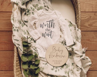 May include: A white baby onesie with the text "worth the wait" in black script. A round wooden sign with the text "Baby Anderson AUGUST 2023" in black script. The onesie and sign are on a white and green patterned blanket in a wicker basket.