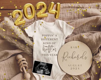 May include: A white onesie with the text "Poppin' a different kind of bottles this year" and a black and white ultrasound image. A wooden sign with the text "Baby Richards August 2024" and gold foil stars are scattered on a brown wooden surface.